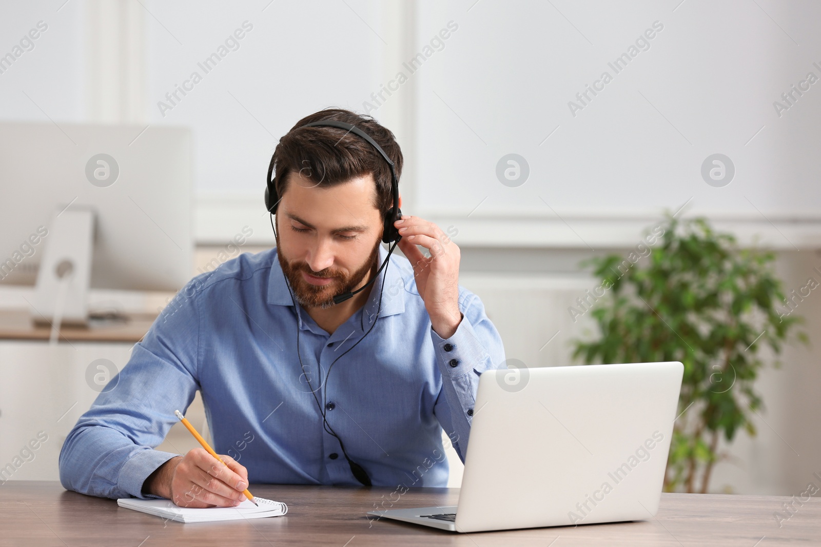 Photo of Hotline operator with headset writing something in notebook while working at wooden table