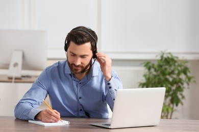 Hotline operator with headset writing something in notebook while working at wooden table