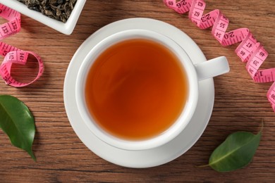 Cup of herbal diet tea and measuring tape on wooden table, flat lay. Weight loss concept