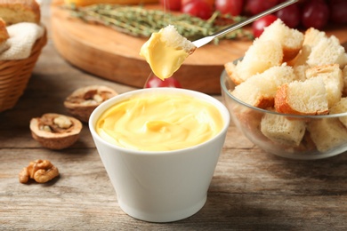 Photo of Piece of bread over bowl with delicious cheese fondue on wooden table