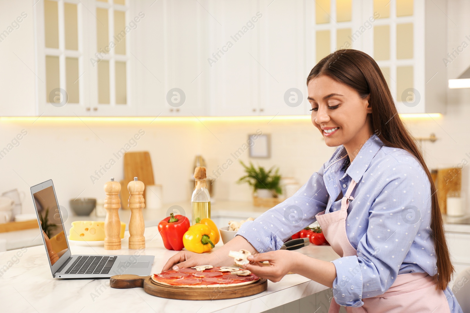 Photo of Woman making pizza while watching online cooking course via laptop in kitchen