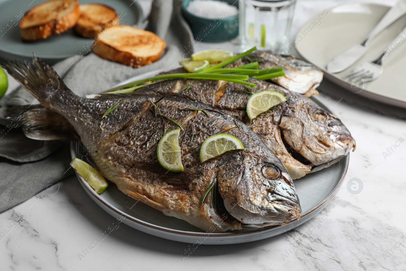 Photo of Seafood. Delicious baked fish served with green onion and lime on white marble table, closeup