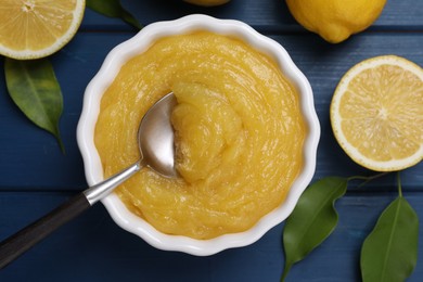 Delicious lemon curd in bowl, fresh citrus fruits and spoon on blue wooden table, flat lay