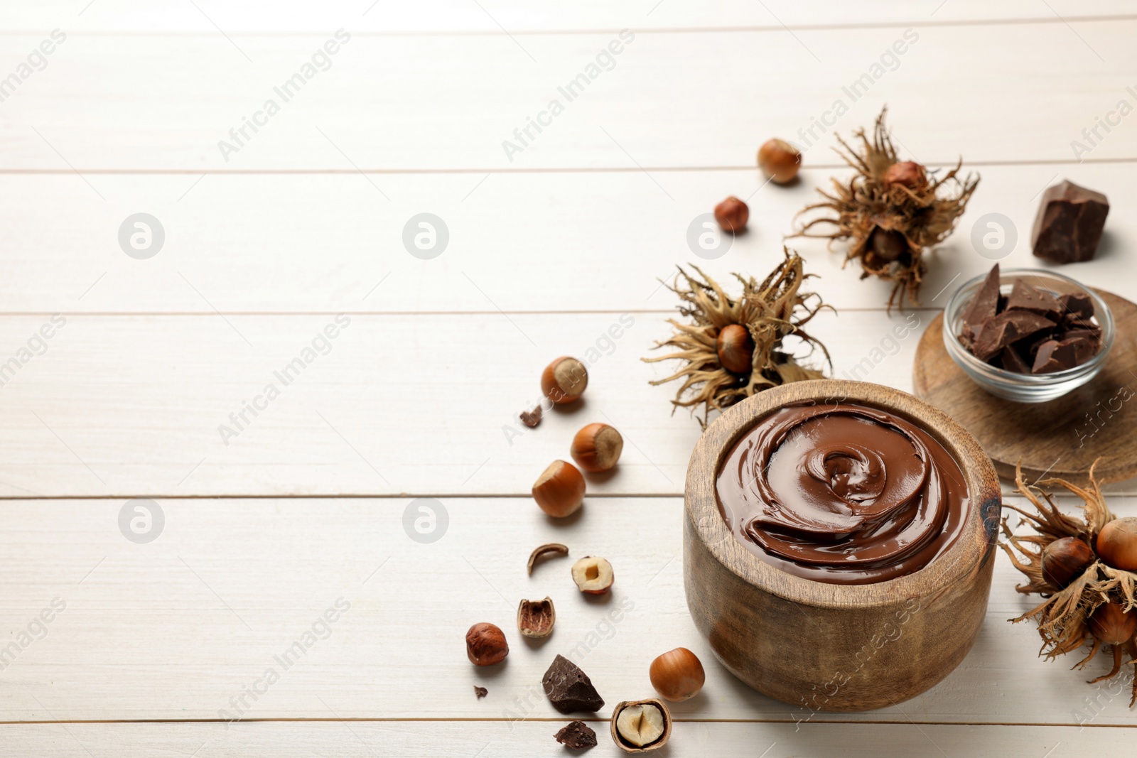 Photo of Bowl of tasty chocolate paste with hazelnuts on white wooden table, space for text