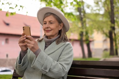 Photo of Beautiful senior woman sitting on bench and using smartphone outdoors, space for text