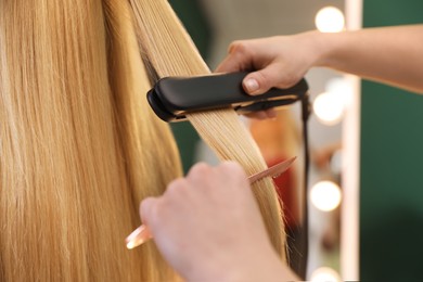 Stylist straightening woman's hair with flat iron in salon, closeup