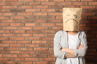 Photo of Woman wearing paper bag with drawn sad face near brick wall. Space for text