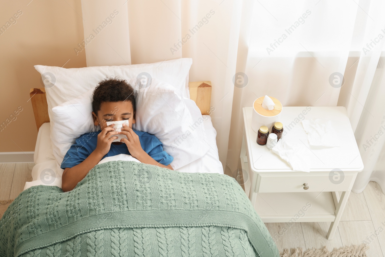 Photo of African-American boy with scarf and tissue blowing nose in bed indoors, above view. Cold symptoms