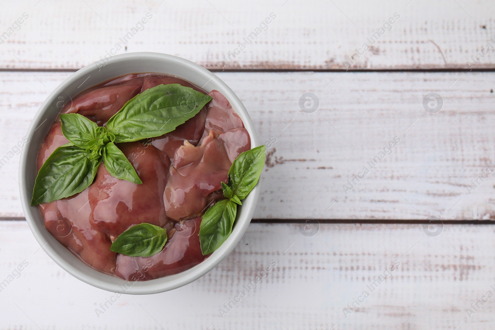 Photo of Bowl with raw chicken liver and basil on white wooden table, top view. Space for text