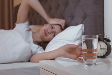 Young woman with terrible headache at night taking glass of water from stand