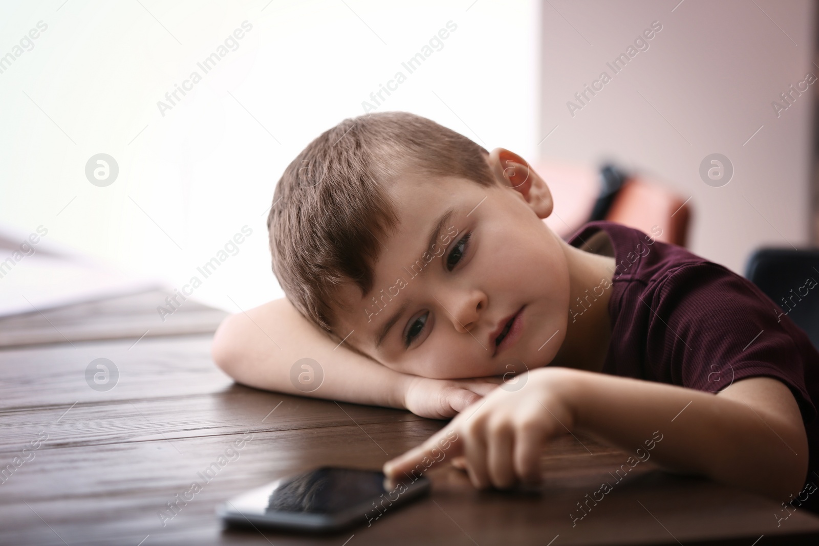 Photo of Sad little boy with mobile phone sitting at table indoors