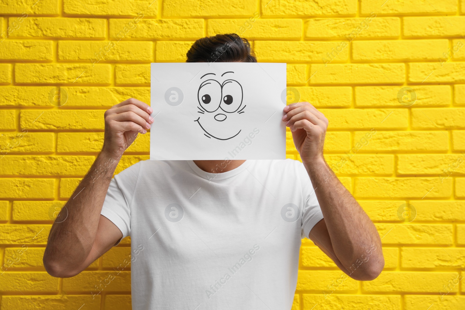 Photo of Man hiding emotions using card with drawn smiling face near yellow brick wall