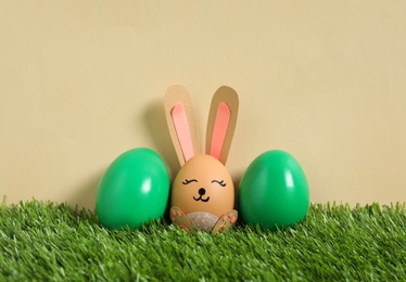 Photo of Bright eggs and brown one as Easter bunny on green grass against beige background