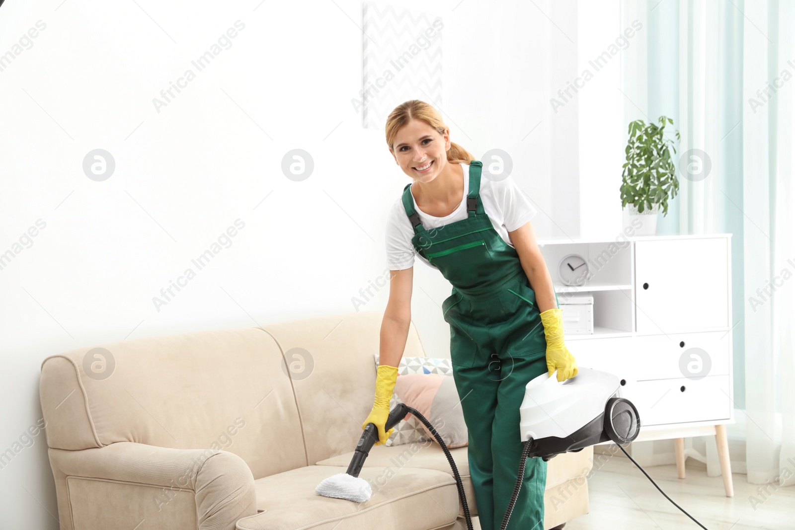 Photo of Female janitor removing dirt from sofa with steam cleaner in room