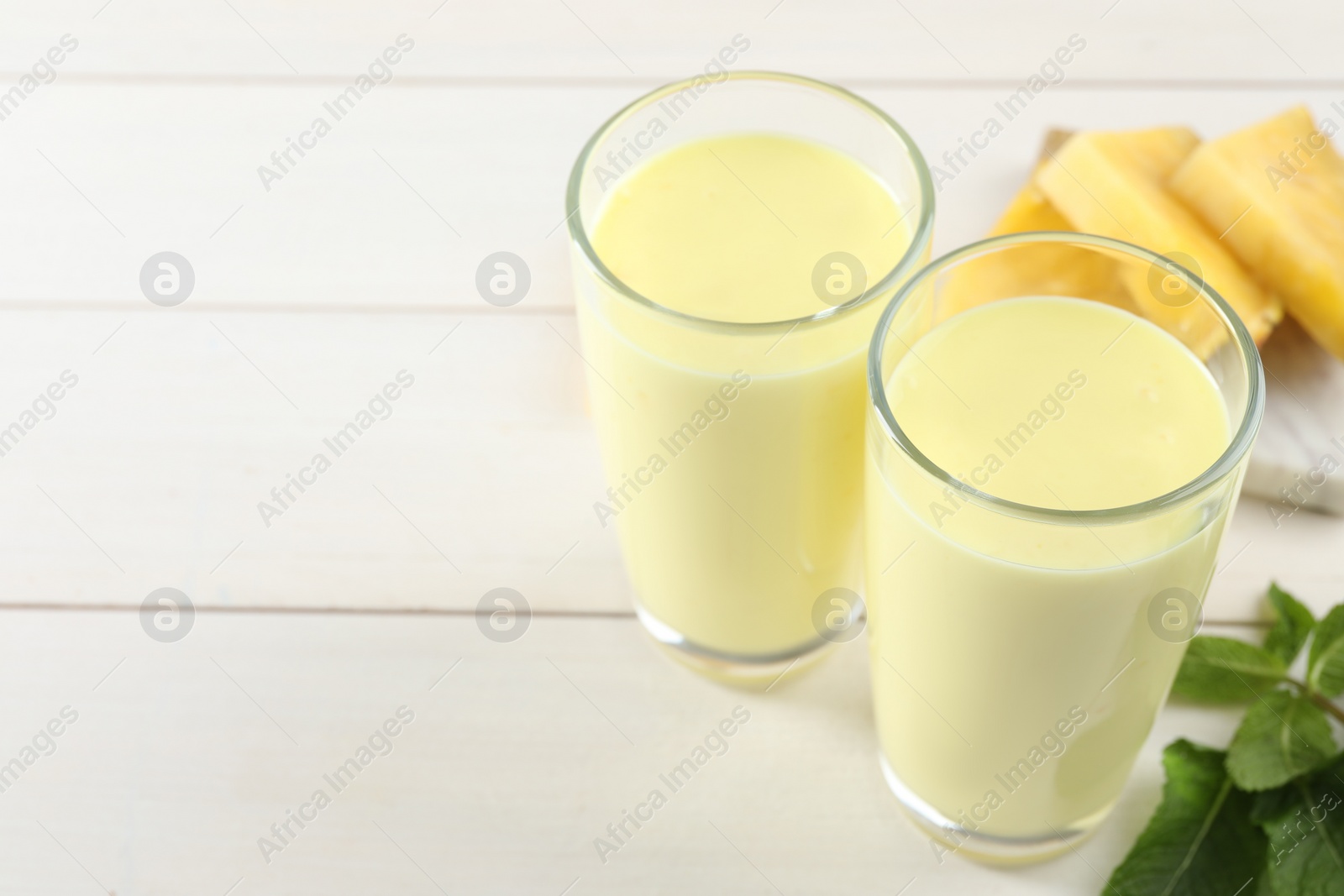 Photo of Glasses of tasty pineapple smoothie, mint and fresh cut fruit on white wooden table. Space for text