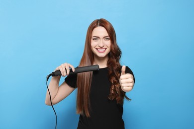 Beautiful woman with hair iron showing thumbs up on light blue background