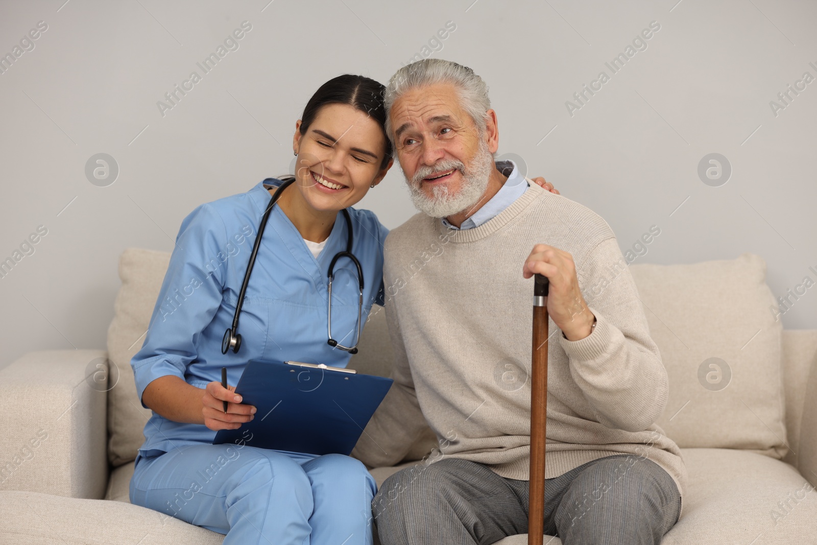 Photo of Health care and support. Nurse holding clipboard and laughing with elderly patient on sofa indoors