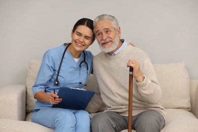 Photo of Health care and support. Nurse holding clipboard and laughing with elderly patient on sofa indoors