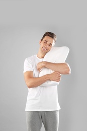 Young man in pajamas embracing pillow on gray background