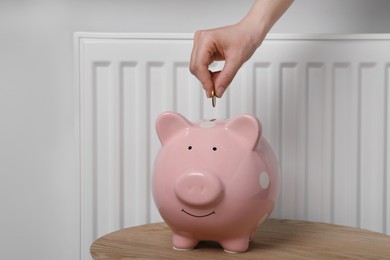 Photo of Woman putting coin into piggy bank near heating radiator, closeup