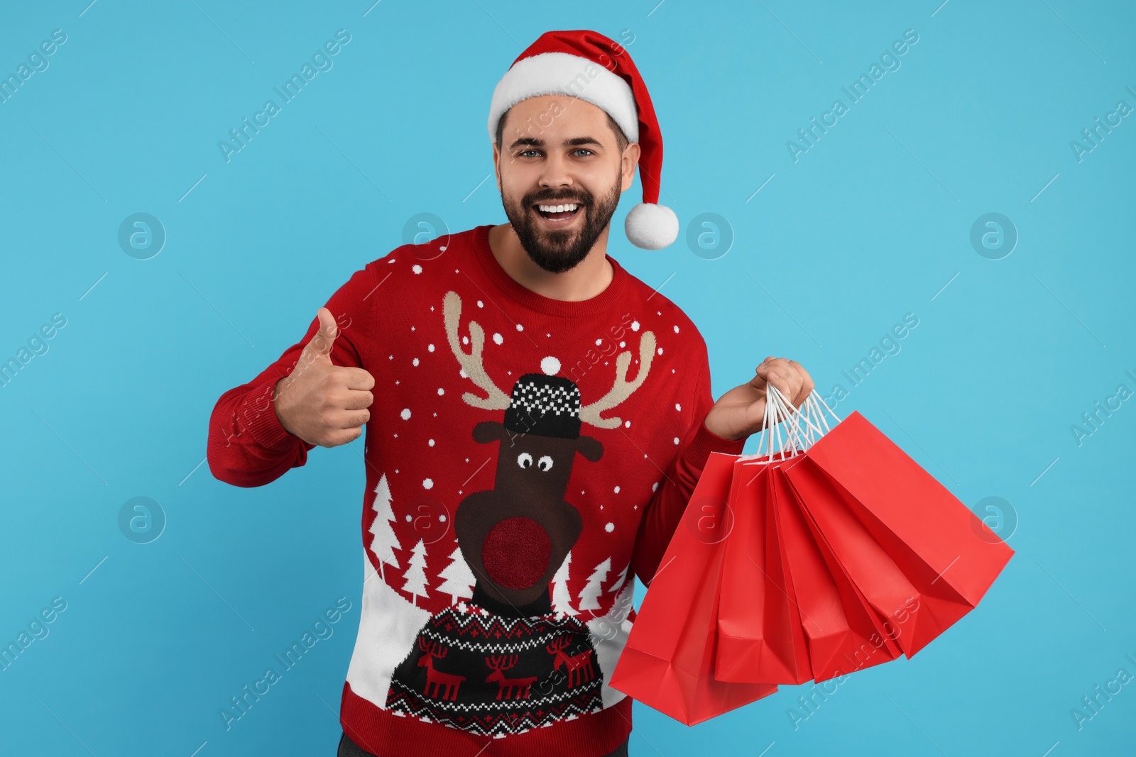 Photo of Happy young man in Christmas sweater and Santa hat with shopping bags on light blue background