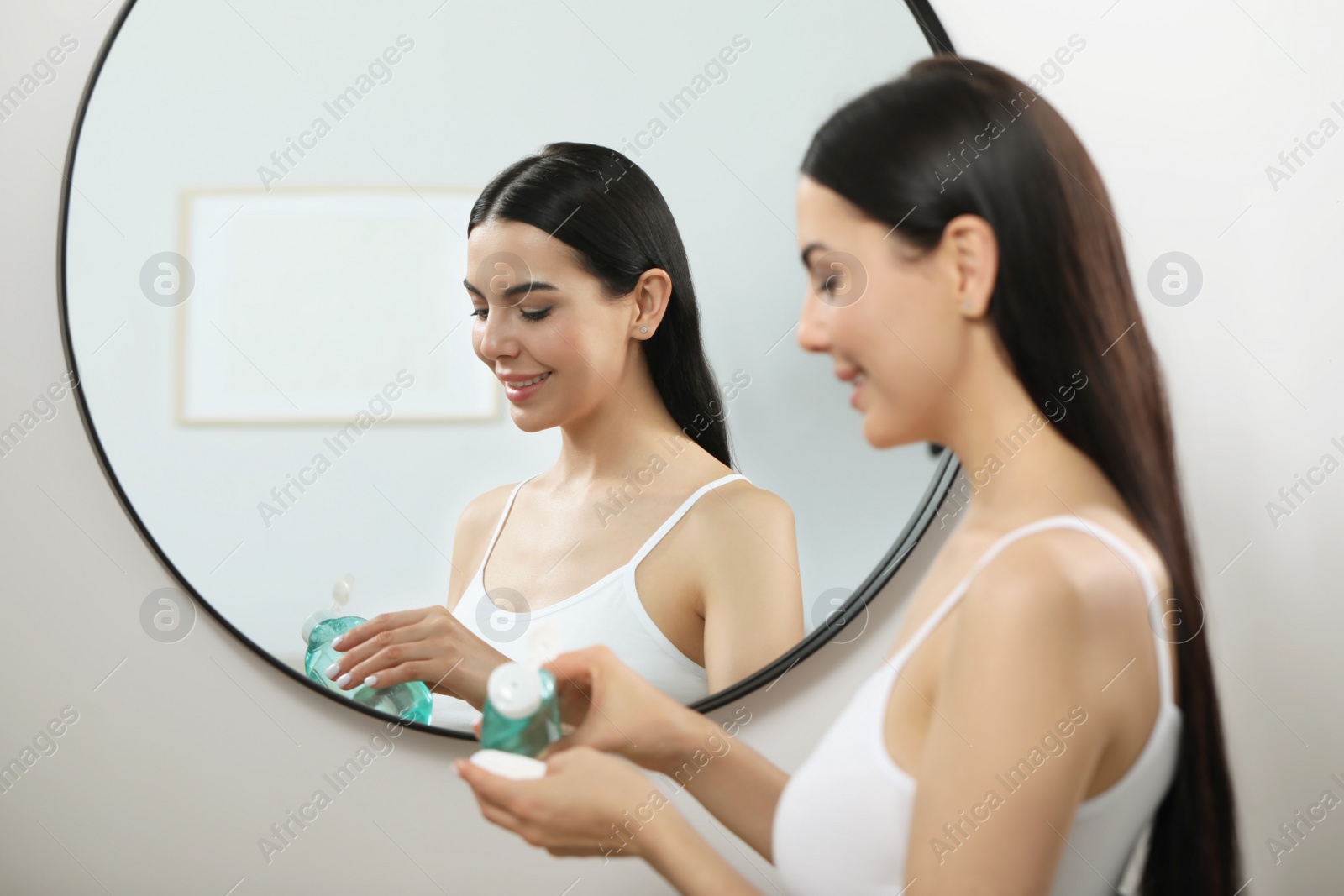 Photo of Young woman pouring micellar water onto cotton pad near mirror indoors