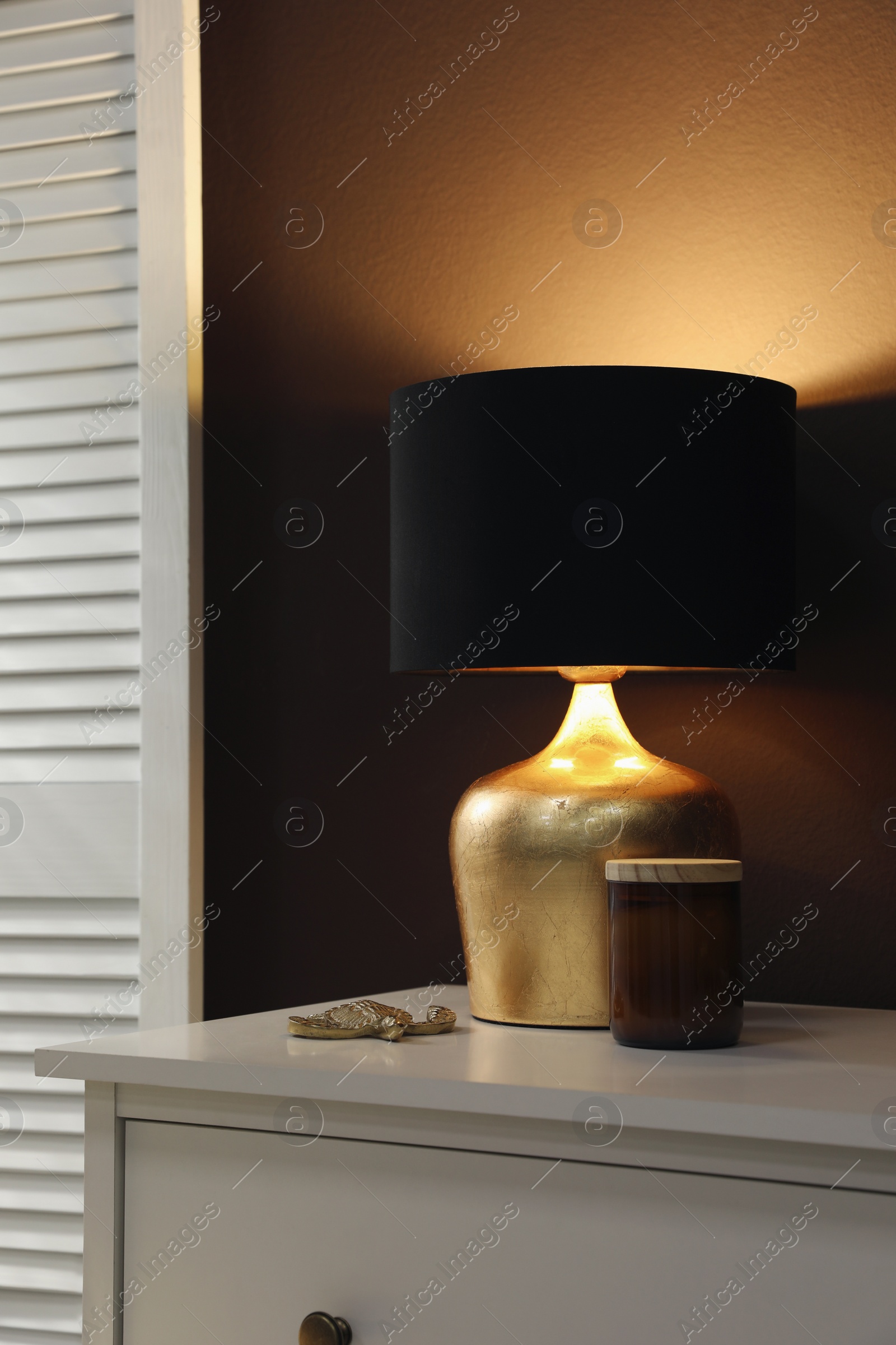 Photo of Stylish lamp and decor on white chest of drawers near brown wall indoors. Interior design