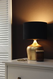 Stylish lamp and decor on white chest of drawers near brown wall indoors. Interior design