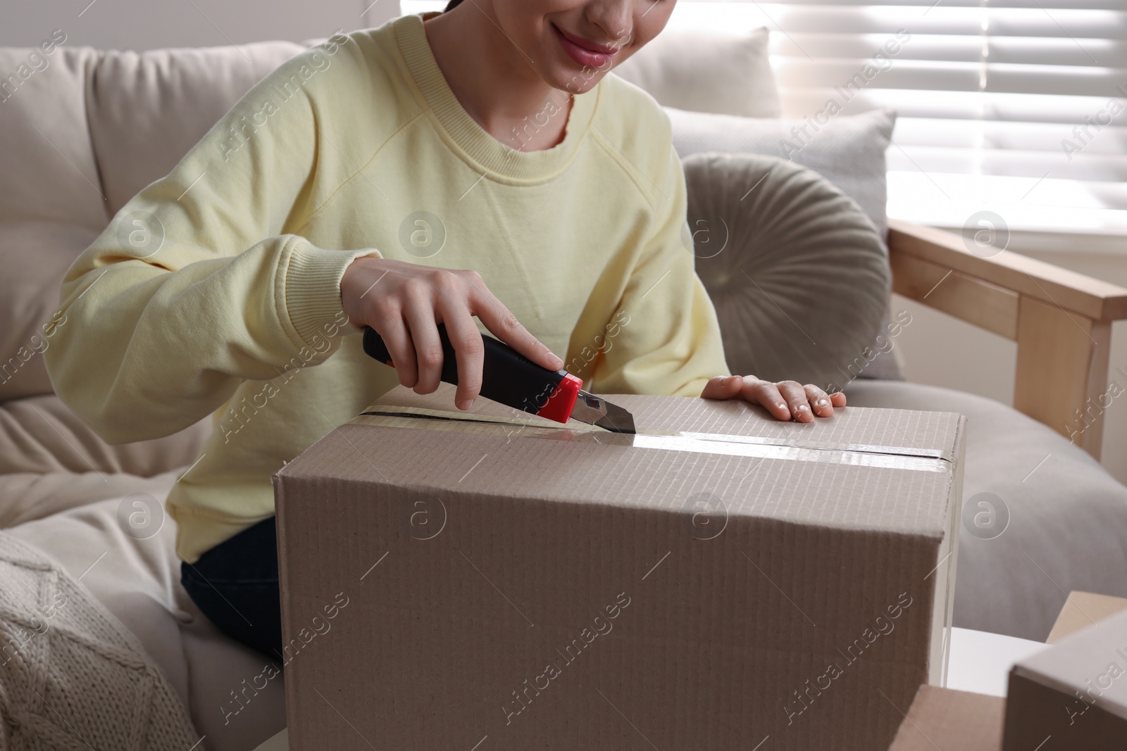 Photo of Young woman using utility knife to open parcel at home, closeup