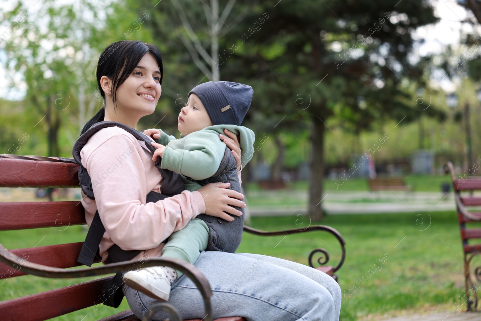 Photo of Mother holding her child in sling (baby carrier) on bench in park. Space for text