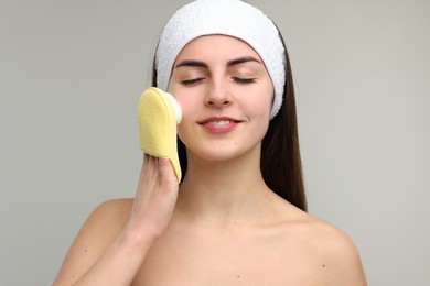 Photo of Young woman with headband washing her face using sponge on light grey background