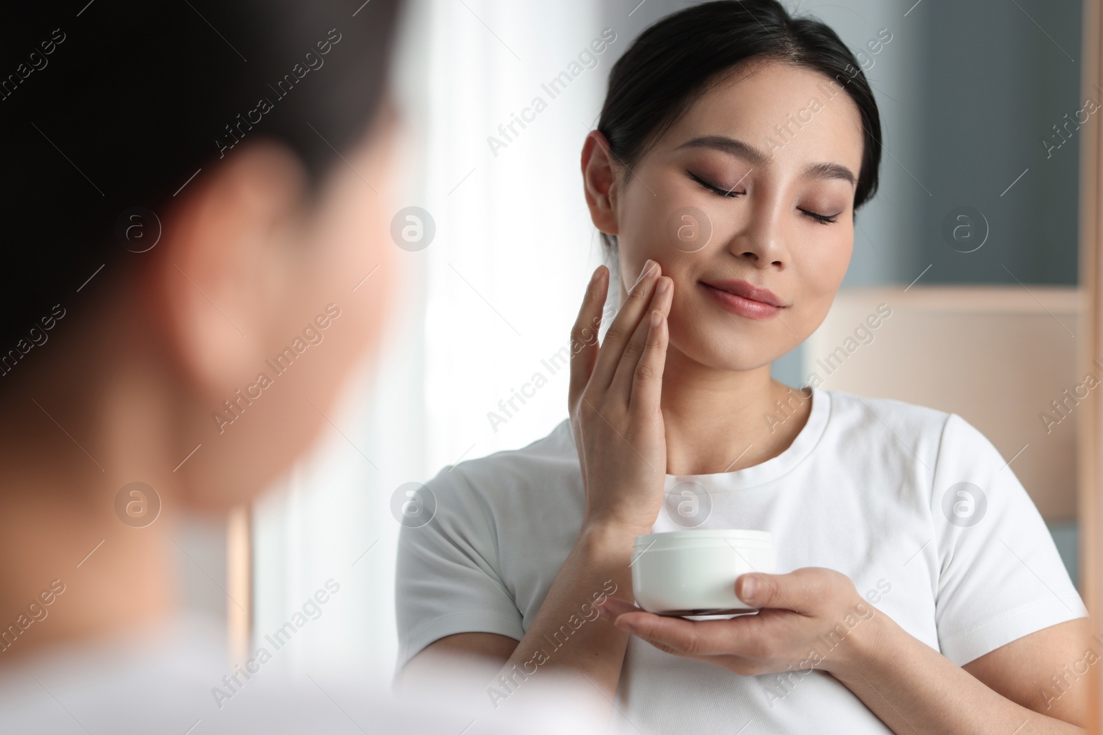 Photo of Happy woman applying face cream near mirror at home