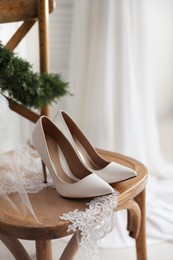 Photo of Pair of white high heel shoes, veil and wreath on wooden chair indoors. Dressing for wedding