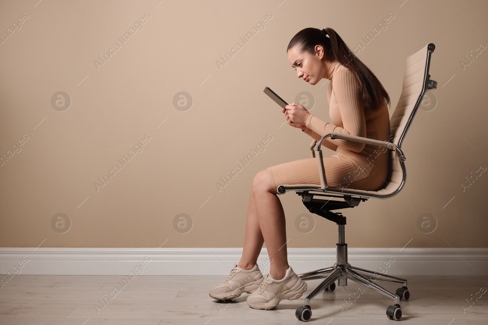 Photo of Young woman with poor posture using tablet while sitting on chair near beige wall, space for text