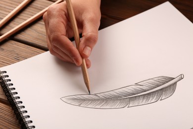 Woman drawing feather with graphite pencil in sketchbook at wooden table, closeup