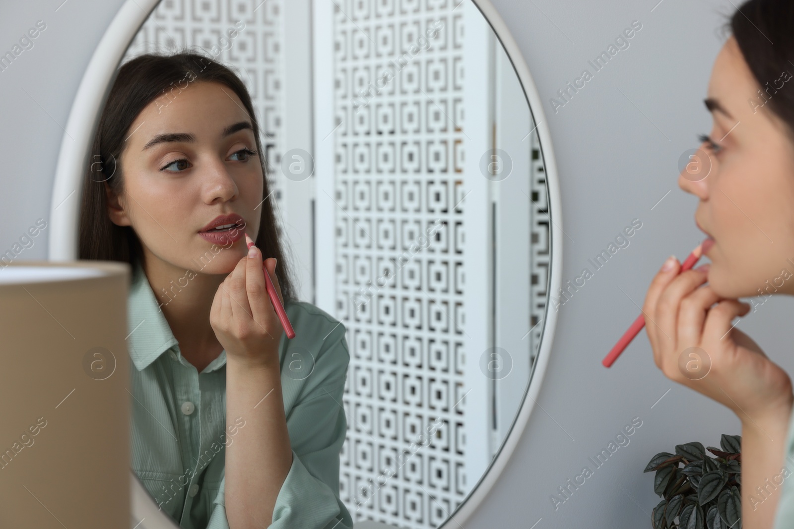 Photo of Beautiful young woman applying cosmetic pencil on lips near mirror indoors