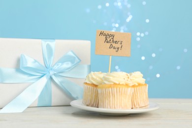 Photo of Happy Father's Day. Tasty cupcakes with greetings and gift box on light wooden table, closeup