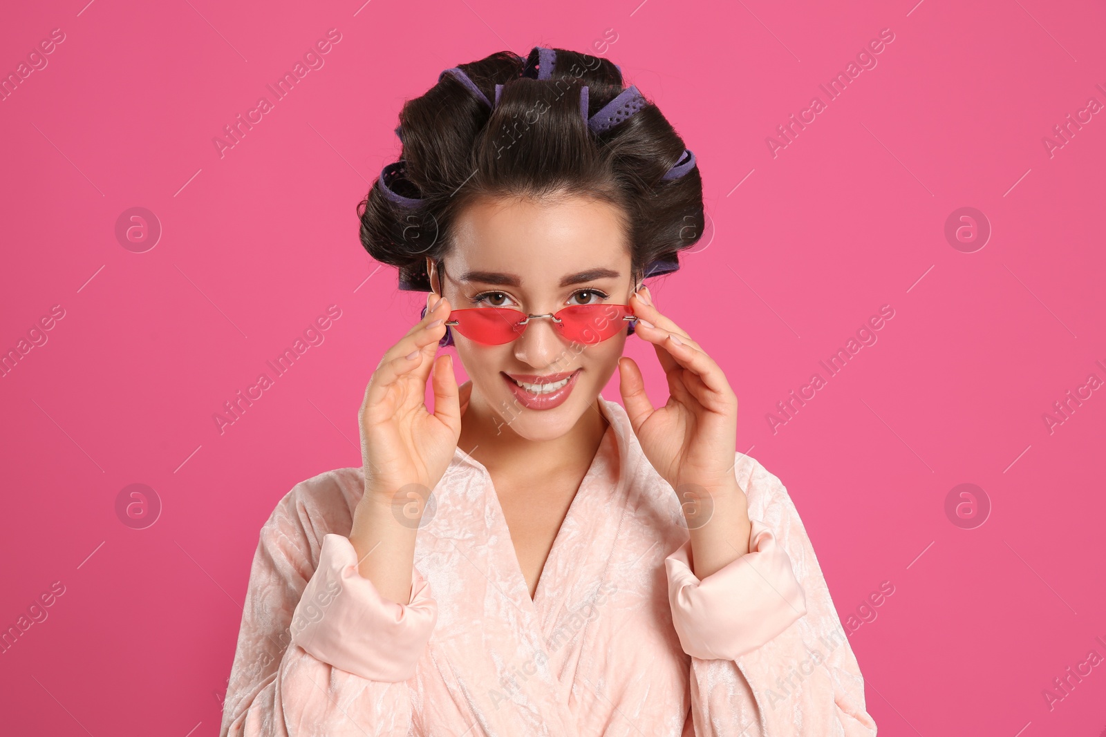 Photo of Happy young woman in bathrobe with hair curlers and sunglasses on pink background