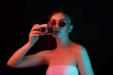 Photo of Beautiful woman with sunglasses and vintage camera posing in neon lights against black background