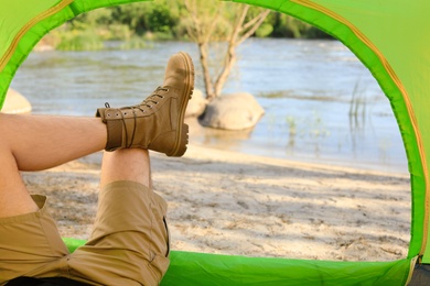 Young man resting in camping tent on riverbank, view from inside