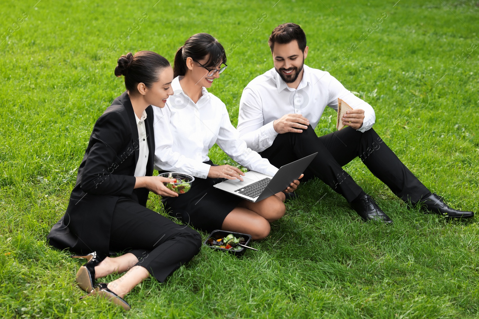 Photo of Happy colleagues with laptop having business lunch on green grass outdoors