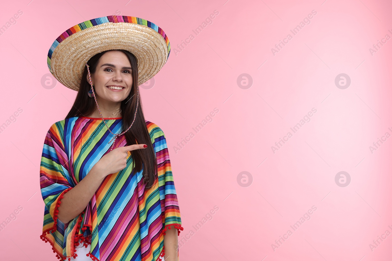 Photo of Young woman in Mexican sombrero hat and poncho pointing at something on pink background. Space for text