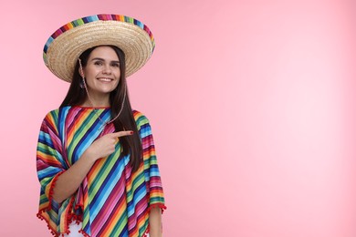 Photo of Young woman in Mexican sombrero hat and poncho pointing at something on pink background. Space for text