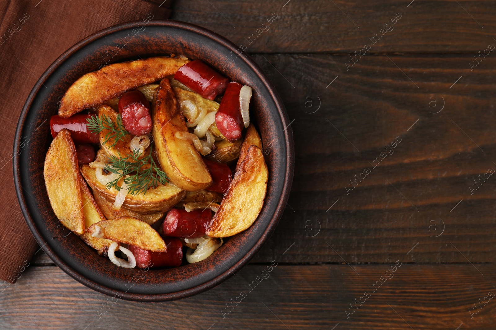 Photo of Delicious baked potato with thin dry smoked sausages, onion and dill in bowl on wooden table, top view. Space for text