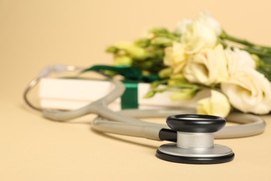 Photo of Stethoscope, gift box and eustoma flowers on beige background, closeup. Happy Doctor's Day