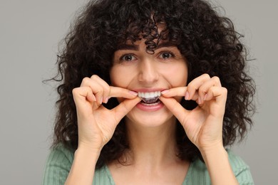 Young woman applying whitening strip on her teeth against grey background