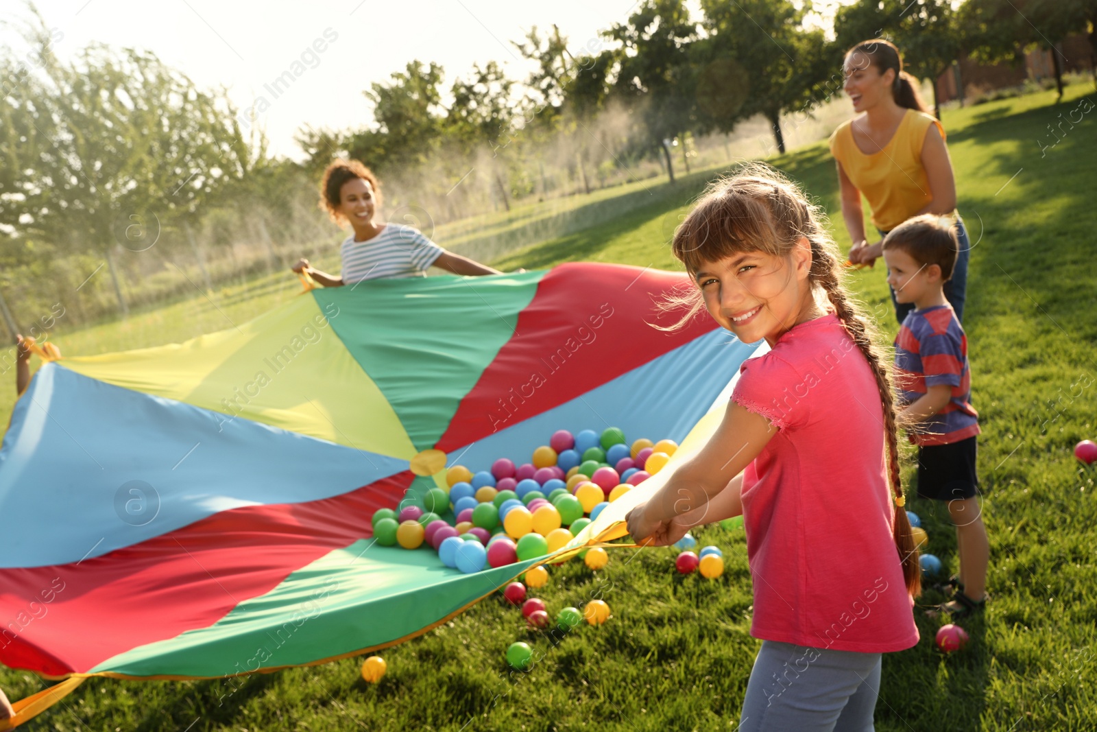 Photo of Group of children and teachers playing with rainbow playground parachute on green grass. Summer camp activity