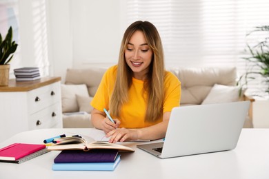 Photo of Young woman writing down notes during webinar at table in room