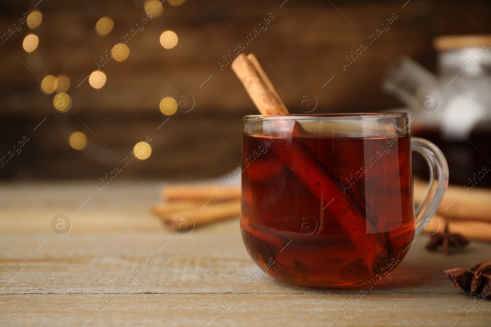 Photo of Glass cup of hot tea with aromatic cinnamon on wooden table against blurred lights. Space for text