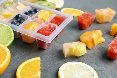 Photo of Ice cubes with fruits and berries in tray near ingredients on grey table